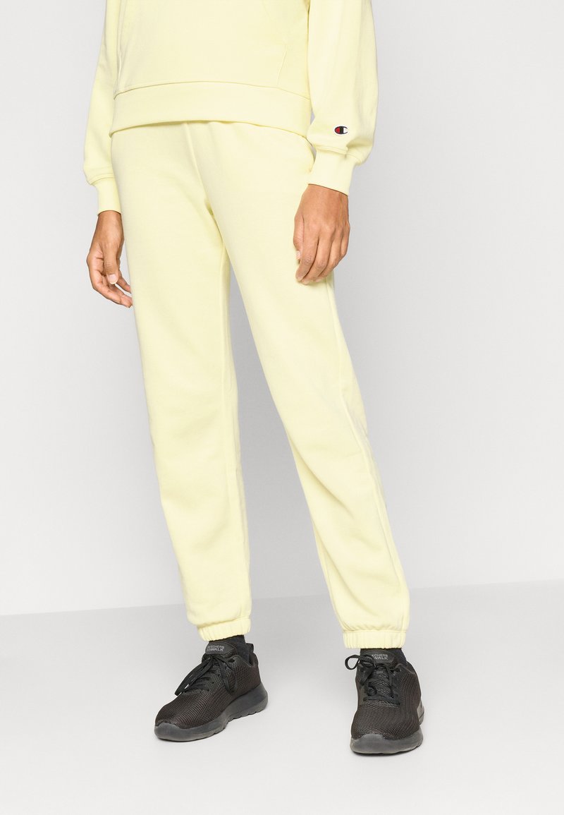 Κίτρινο φούτερ και αντίστοιχο παντελόνι φόρμας από μαλακό ύφασμα, με ελαστικές μανσέτες και λογότυπο στο μανίκι. Φοριέται με μαύρα αθλητικά παπούτσια.