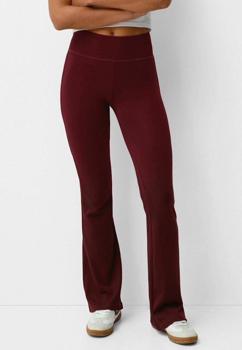Personne portant un pantalon évasé bordeaux taille haute et des baskets blanches, debout les bras croisés devant un fond blanc.