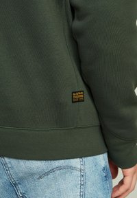 Sweatshirt de color verde oscuro con dobladillo y puños acanalados, que presenta una pequeña etiqueta de marca negra y amarilla en el lado cerca de la costura inferior.