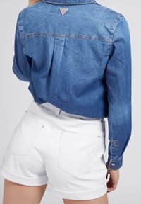 La giacca di jeans presenta un colore azzurro chiaro con una texture leggermente stropicciata e un piccolo patch con il logo. Abbinata a pantaloni corti bianchi arrotolati.