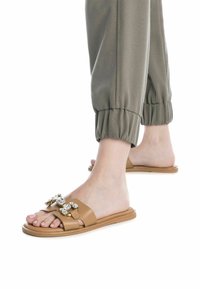 Chanclas marrones con suela plana, decoradas con gemas transparentes y diseño de punta abierta. Combinadas con pantalones de puño elástico en verde oliva.