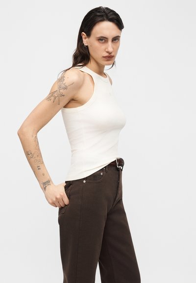 Γυναίκα με σκούρα μαλλιά που φοράει λευκό αμάνικο τοπ και σκούρο παντελόνι, εμφανίζοντας τα τατουάζ στο αριστερό της χέρι μπροστά από ένα απλό φόντο.