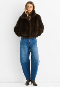 Young woman wearing a dark brown fur jacket, blue high-waisted jeans, and brown heeled ankle boots standing against a white background.