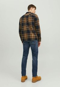 Camisa de cuadros en marrón, azul marino y beige; combinada con jeans azules y botas de color beige. La camisa tiene un cuello clásico y un detalle en el yugo trasero.