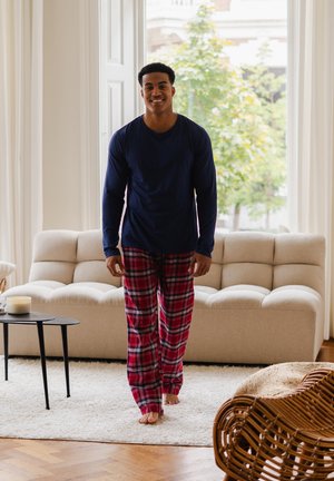 Chemise à manches longues marine, pantalon de pyjama à carreaux rouges. Tissu lisse, coupe décontractée. Tapis clair et canapé beige visibles dans une pièce lumineuse.