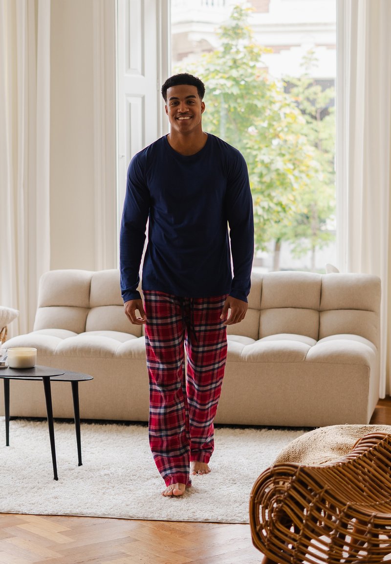 Camisa de manga larga de marina, pantalones de pijama rojos a cuadros. Tejido suave, corte relajado. Alfombra ligera y sofá beige visibles en una habitación luminosa.