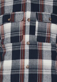 Camicia in flanella a quadri con colori blu navy, bianco e rosso. Due tasche sul petto con chiusura a bottone e bottoni marroni sulla parte anteriore.
