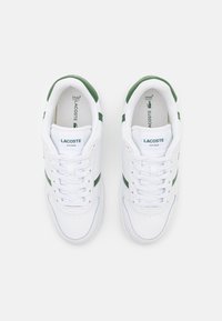 White sneakers with green accents feature a smooth leather upper, round toe, and flat laces. Branding is displayed on the tongue and heel.