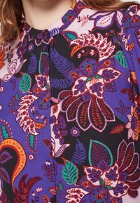 Blusa a fiori viola con dettagli intricati in rosso, verde e bianco. Presenta un collo arrotondato e chiusura a bottoni sul davanti.
