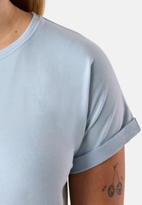 T-shirt azzurro chiaro con maniche corte e risvolti, realizzato in tessuto morbido. Presenta un collo a giro e una texture sottile. Tatuaggio visibile sul braccio.