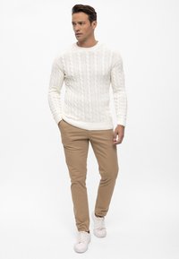 Suéter blanco de punto trenzado con puños acanalados, combinado con pantalones beige claros y zapatillas blancas. Modelo de pie con expresión neutral.