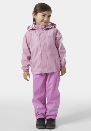 Mladá dívka stojí a usmívá se, má na sobě růžovou voděodolnou bundu a kalhoty s reflexními pruhy a černé boty na bílém pozadí.