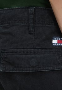 Κοντινή λήψη τσέπης μαύρου παντελονιού με καπάκι και μικρό λογότυπο Tommy Jeans στη δεξιά πλευρά.