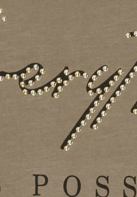 Beige stof med sort kursiv tekst dekoreret med guld nitter, hvilket skaber et struktureret design. Overfladen er glat med en let glans.