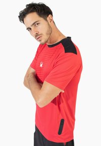 Camiseta atlética roja con un acento negro en el hombro, patrón texturizado en la parte frontal y un bolsillo con cremallera en el lateral.
