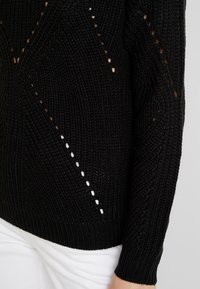 Nahaufnahme eines schwarzen Strickpullovers mit einem strukturierten Muster und offenen Maschen, kombiniert mit weißen Hosen. Lässiges Modedetail.