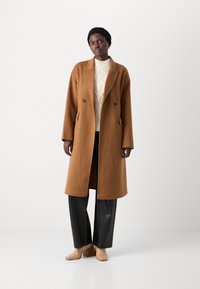 Brauner Wollmischungs-Mantel mit einem langen, lockeren Schnitt, doppelreihigem Design, großen Revers und seitlichen Taschen, getragen über einem cremefarbenen Pullover und schwarzen Hosen.
