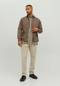 Brun knap-jacket over en ternet skjorte, beige bukser og mørke sko. Jakken har en brystlomme og knapmanchetter. Glatte teksturer.