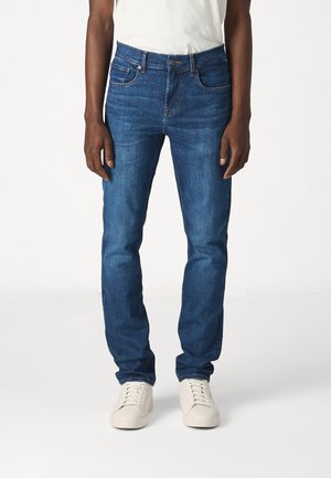 Dunkelblaue Denim-Jeans mit schlankem Schnitt, ausgestattet mit klassischem Fünf-Taschen-Design und dezenten Nähten. Kombiniert mit weißen Sneakers.