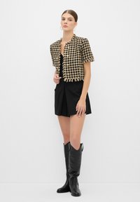 Houndstooth kort jacka i svart och beige, med korta ärmar och fransig nederkant, tillsammans med en svart klänning och knähöga svarta stövlar.