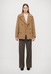 Podwójnie zapięty, brązowy płaszcz o puszystej fakturze z dużymi brązowymi guzikami i klapami, zestawiony z luźnymi, szarymi spodniami.