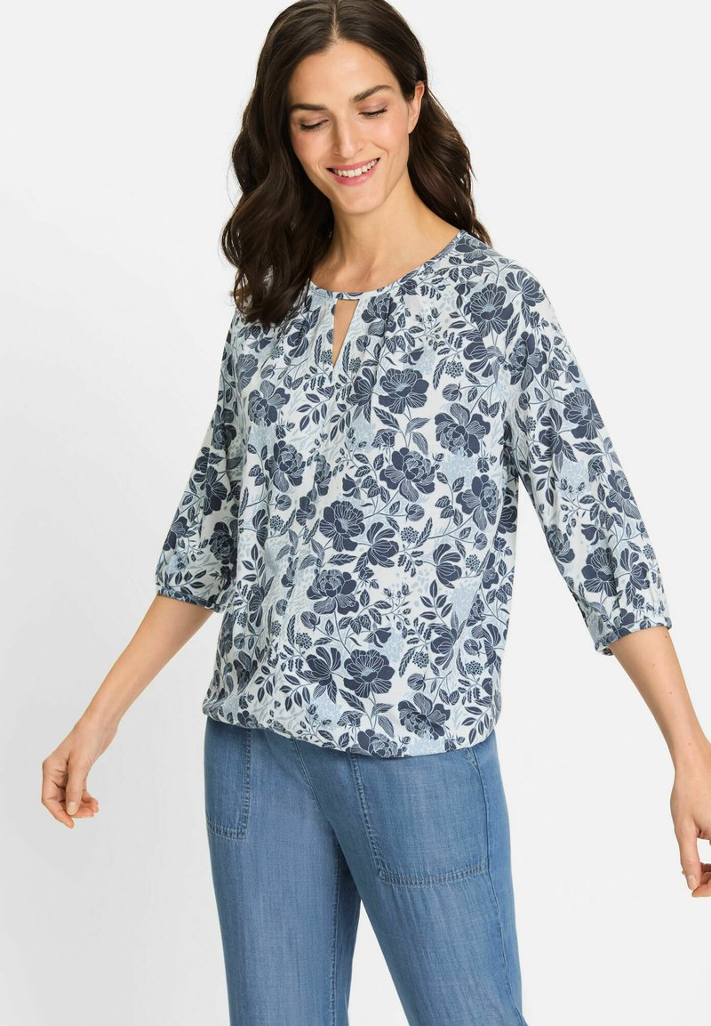 Hellbluse mit floralem Muster, die marineblaue Blumen und Blätter zeigt, kurze elastische Ärmel und einen Schlüsselloch-Ausschnitt hat. Kombiniert mit hellblauen Jeans.
