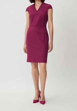 Femme portant une robe magenta sans manches, longueur genou, avec un col en V, associée à des chaussures à talons hauts magenta coordonnées, debout devant un fond uni.