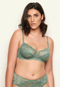 Smaragdgrön spets-bh med mjuka kupor och justerbara axelband, med en vågig kant och blommönster. Matchande spets-trosor nedanför.