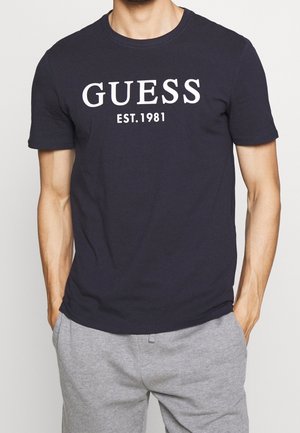 Mężczyzna noszący czarną koszulkę Guess z białym tekstem "GUESS EST. 1981" i jasnoszare dresy, ręce w kieszeniach.