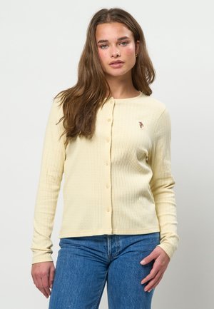 Junge Frau mit langen braunen Haaren, die eine hellgelbe, geknöpfte Strickjacke und blaue Jeans trägt und vor einem schlichten hellen Hintergrund steht.