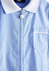 Camicia a quadri blu e bianca con colletto bianco, cursore della zip a forma di fiore e piccola tasca frontale con bordatura in pizzo.