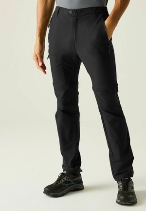 Uomo che indossa pantaloni da trekking neri con zip staccabile e scarpe da trekking nere, in piedi con la mano sinistra in tasca su sfondo semplice.