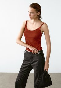 Rostfarbener gerippter Tank-Top mit dünnen Trägern, kombiniert mit hoch taillierten schwarzen Hosen mit silbernen Schnallenakzenten. Das Model hält eine schwarze Clutch.