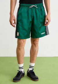 Zelené saténové sportovní šortky s černými bočními pruhy, elastickým pasem a šňůrkou, se znakem a detailem nášivky Adidas v bílé barvě.