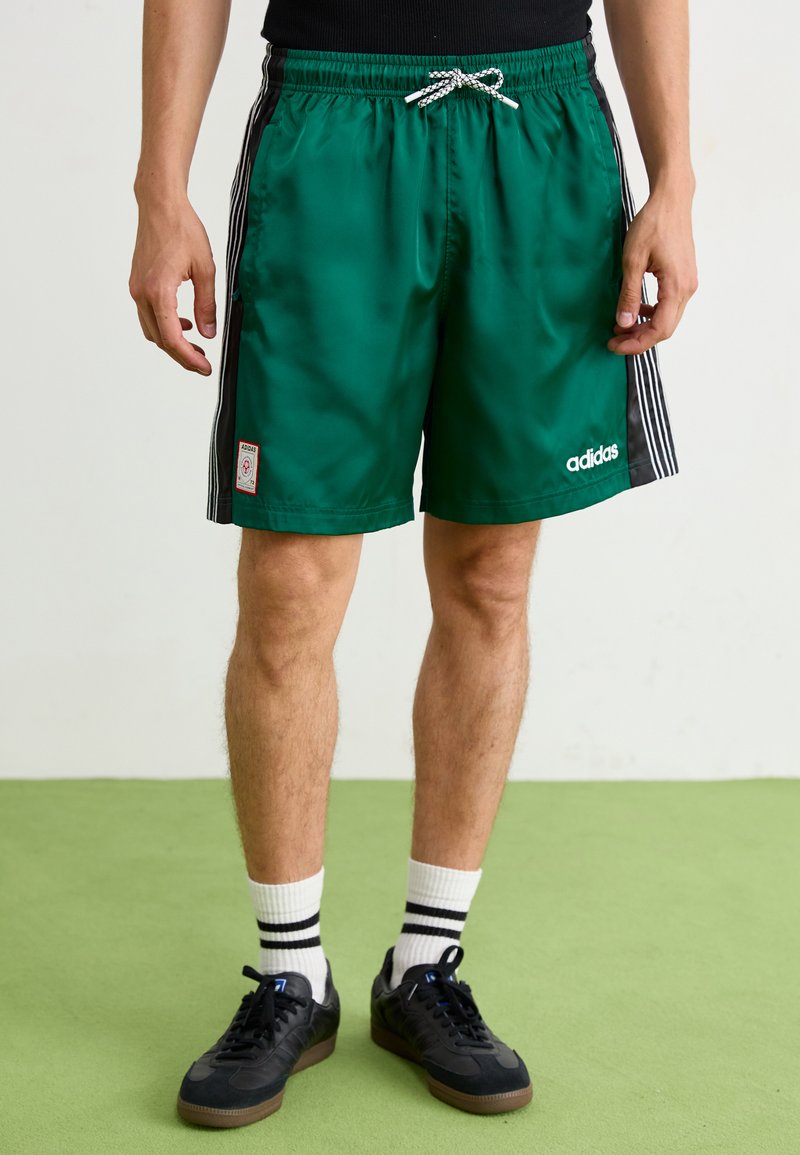 Zelené saténové sportovní šortky s černými bočními pruhy, elastickým pasem a šňůrkou, se znakem a detailem nášivky Adidas v bílé barvě.