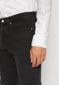 Hombre vistiendo jeans negros y una camisa de mangas largas blanca, con la mano apoyada sobre la cadera, frente a un fondo claro.