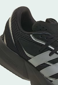 Čierna športová topánka s vrchnou časťou z sieťoviny, syntetickými doplnkami a hrubou podrážkou. Strieborné prúžky prechádzajú po stranách a zdôrazňujú dizajn.