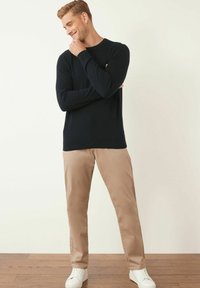 Marineblauer langärmeliger Pullover kombiniert mit beigen Chinos und weißen Sneakers. Der Pullover hat einen Rundhalsausschnitt und gerippte Bündchen. Hintergrund aus Holzboden.
