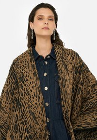 Hnedo-čierny leopardí vzorovaný šál z mäkkej tkaniny, prehodený cez tmavú džínovú košeľu s kovovými gombíkmi.