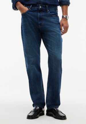 Homme portant un jean bleu foncé, des mocassins en cuir noir, une chemise bleu foncé, avec une main dans la poche et une montre-bracelet à l'autre poignet.