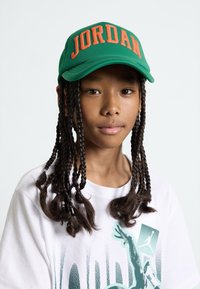 Gorra de béisbol verde con letras naranjas "JORDAN", combinada con una camiseta blanca con un estampado gráfico, sobre una persona con cabello trenzado.