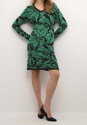 Kobieta ubrana w zieloną i czarną sukienkę w kwiaty do kolan z długimi rękawami oraz czarne szpilki, stojąca na tle gładkiej ściany.