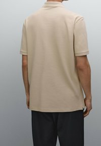 Beige kortärmad polotröja med krage och subtilt strukturerat mönster, designad med sidoklipp i fållen och en slät yta.