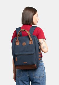 Zaino in tessuto navy con dettagli in pelle marrone, manico superiore e spallacci regolabili. Presenta una tasca frontale con zip e una patch con logo.