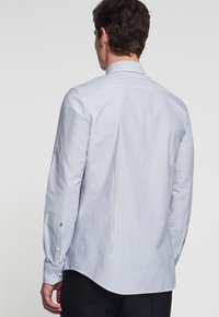 Man die met zijn rug naar de camera staat, gekleed in een wit shirt met dunne verticale blauwe strepen en een zwarte broek, tegen een effen lichte achtergrond.