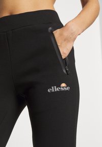 Schwarze Jogginghosen mit geripptem Bund, seitlicher Reißverschlusstasche und kleinem "ellesse"-Logo in Grautönen und farblichen Akzenten. Weiche Textur, figurbetontes Design.