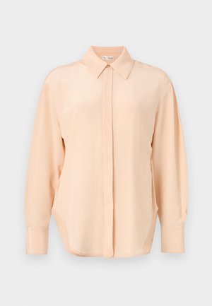 Femeie cu păr închis la culoare, purtând o bluză de culoarea piersicii cu mânecă lungă și pantaloni negri largi, stând în fața unui fundal simplu, luminos.