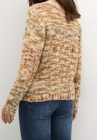 Pletený svetr v béžové barvě s vícebarevnými skvrnami; volnější střih, dlouhé rukávy, žebrované manžety a lem, textura působí měkce a teple.