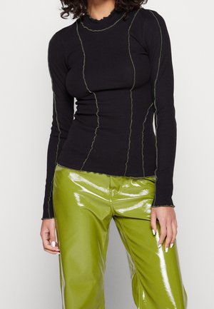 Femme portant un haut noir cintré à manches longues avec des détails de coutures vertes et un pantalon en vinyle vert citron brillant sur un fond uni.