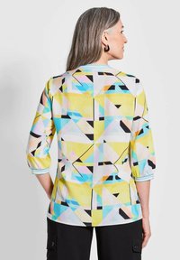Bunt gemusterte Bluse mit geometrischen Motiven in Gelb, Blau, Pink und Schwarz. Leichter Stoff mit 3/4-Ärmeln und blauen Streifen am Saum.
